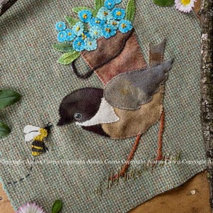 May include: A textile art piece featuring a bird carrying a brown bag filled with blue flowers. A yellow and black bee hovers near the bird. The background is a textured green fabric, with a daisy and other floral elements.