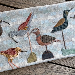 May include: A decorative textile with six bird figures in brown, blue, and red. Each bird is perched on a thin stem with a square or round base. The background has a light blue and white striped pattern. The textile is likely for home decor.