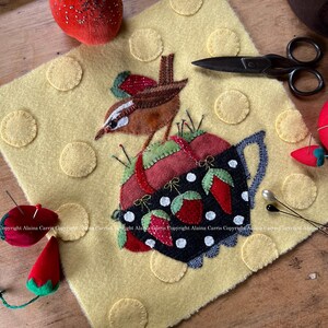 May include: A felt applique of a brown bird perched on a black and white teacup filled with red strawberries. The teacup is surrounded by yellow felt circles. The background is a yellow felt fabric.