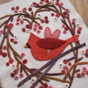 Puede incluir: Un pájaro cardenal rojo con una rama marrón y bayas rojas, bordado sobre un fondo de tela blanca. El diseño está rodeado por una corona de ramas marrones con bayas rojas y flores blancas.