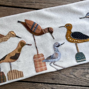 May include: A white fabric with six embroidered birds, each with a different colour and pattern. The birds are standing on wooden bases.