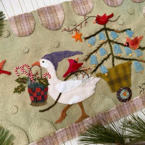 May include: A whimsical Christmas-themed felt table runner. It features a goose wearing a hat, carrying a basket of candy canes and holly. A Christmas tree with blue bell ornaments and red cardinals are also depicted. The runner has a light green background with decorative stitching.