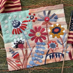 May include: A patriotic textile art piece featuring stylized flowers and American flags. The artwork incorporates red, white, and blue colors with star and stripe motifs. The piece is likely handmade, with a rustic, folk art aesthetic.