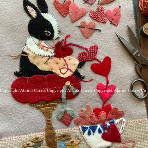 May include: A black and white bunny sits on a red cushion, sewing a string of red hearts. The bunny is wearing a white collar and a pink bow. There are spools of thread and a cup of hearts on a table in the background. The text "Copyright Alaina Carris Copyright Alaina Carris Copyright Alaina Carris Co" is at the bottom of the image.