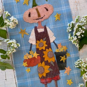 May include: A textile wall hanging featuring a felted pumpkin-headed figure wearing a brown striped apron with sunflower appliqués. The figure holds a red basket and a dark bucket filled with yellow stars on a light blue plaid fabric background.