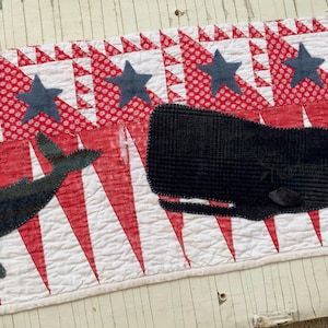 May include: A rectangular fabric quilt runner featuring a white background with red triangular patterns and dark blue star appliques. Two dark whales are appliqued onto the surface, and the text Avery Hill is visible in the bottom right corner.