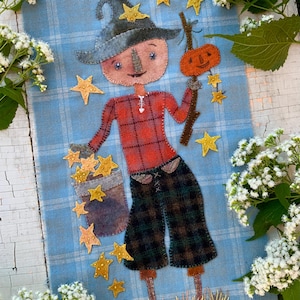 May include: A fabric wall hanging on a light blue plaid background featuring a stitched scarecrow figure wearing a hat, a red plaid shirt, and dark plaid pants. The figure holds a pumpkin on a stick and a bucket filled with yellow stars.