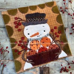 May include: A handcrafted felt snowman with a carrot nose, wearing a hat and scarf, holding a basket of autumn leaves and pumpkins. The artwork is set against a patterned background with decorative stitching and red berry accents, evoking a cozy, seasonal theme.