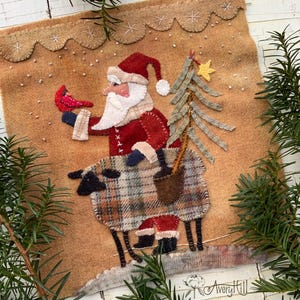 Puede incluir: Aplicación de fieltro navideña hecha a mano que representa a Papá Noel montado en una oveja. Papá Noel viste un traje y gorro rojos, sosteniendo un pájaro rojo y un pequeño árbol de Navidad. El fondo es beige con costuras decorativas.