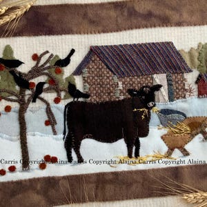 Può includere: Un arazzo da parete in feltro applicato con una mucca marrone in piedi davanti a un fienile marrone con tetto rosso. C'è un albero con uccelli neri e mele rosse. Lo sfondo è bianco con un bordo marrone.