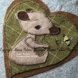 May include: A handmade felt heart-shaped applique featuring a sleeping bear and a bee. The bear is light brown with dark brown accents, stitched onto a green plaid background. Finished size is 10" x 10".