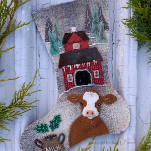 Puede incluir: Una media de fieltro gris con un granero rojo, una vaca marrón y una jarra de leche verde. La media está decorada con árboles de fieltro verde y un banco de nieve de fieltro blanco. La media tiene el texto "Cooper's Cow" y "2024 Stocking".