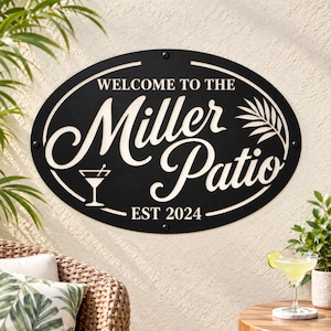Op de afbeelding: Een ovaal zwart metalen bord met witte letters met de tekst "WELCOME TO THE Miller Patio EST 2024". Het bord bevat een afbeelding van een martiniglas en een palmblad en is bevestigd met vier zichtbare schroeven aan de boven-, onder- en zijkanten.