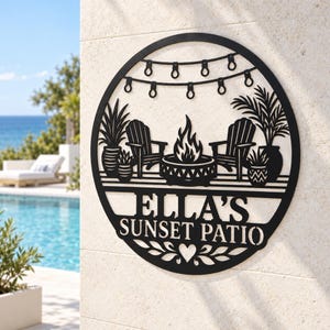 May include: A circular black metal wall sign featuring a cutout design of two chairs, a fire pit, potted plants, and string lights. The text ELLA'S SUNSET PATIO is centred in the middle, with a small heart icon below the lettering.