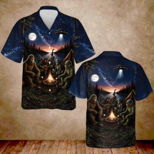 May include: A short-sleeve button-down shirt featuring a dark night sky print with a campfire scene. The graphic depicts Bigfoot, a moth-like creature, an alien, and a swamp monster roasting marshmallows near a lake under a UFO and a full moon.