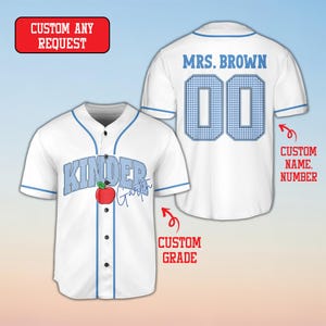 May include: A white baseball jersey with blue trim, featuring the text KINDER with a red apple graphic and the script word Garten on the front. The back displays the text MRS. BROWN and the number 00 in a blue and white checkered pattern.