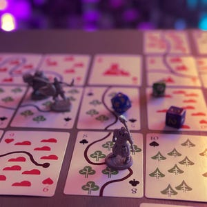 May include: A tabletop board game featuring a grid of playing cards with red heart and green tree symbols, connected by dark lines. Two grey miniature warrior figures stand on the cards, accompanied by a blue and a purple multi-sided die.