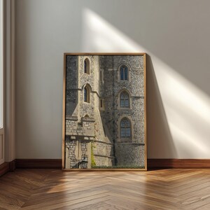 May include: A framed print of a stone castle with arched windows. The castle walls are made of gray stone, and the windows have blue glass. The frame is made of wood, and the print is displayed on a white wall above a wooden floor.
