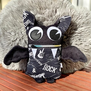 May include: A black plush monster with large white eyes, bat wings, and a zipper mouth. The monster's body is decorated with a black and white rock and roll themed fabric that says "Forever Rock" and features a guitar graphic.