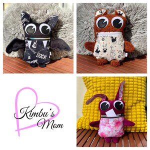 Può includere: Tre peluche fatti a mano: un pipistrello con ali nere e una tasca a tema rock, una creatura marrone simile a un orso con una tasca fantasia e un coniglio rosa con una tasca floreale. I giocattoli hanno grandi occhi espressivi.