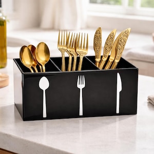 Könnte beinhalten: Ein schwarzer Besteckhalter mit weißen Umrissen von Löffel, Gabel und Messer. Der Halter enthält goldfarbene Löffel, Gabeln und Messer. Das Besteck steht auf einer weißen Arbeitsplatte.
