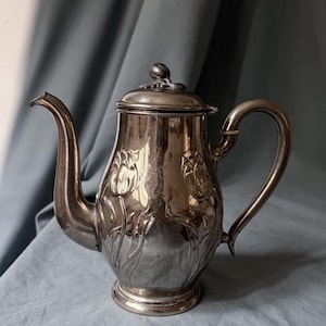 May include: A silver teapot with a curved spout and handle. The teapot has embossed floral patterns and a lid with a small knob. The teapot is likely made of metal and sits on a light blue surface.