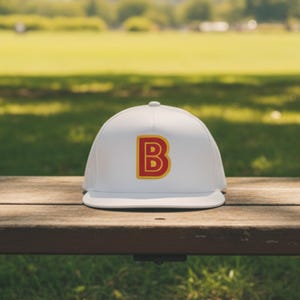 May include: A white baseball cap with a flat brim, featuring a large, stylized letter "B" in red with a gold outline. The cap is resting on a wooden bench, with a blurred green park background.