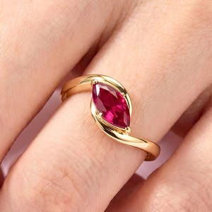 May include: A gold ring with a marquise-cut, deep red gemstone. The ring's band curves around the stone, creating a modern design. The ring is shown on a finger against a soft pink background.
