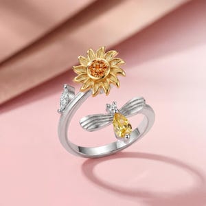 Puede incluir: Anillo plateado con diseño de girasol y abeja. El girasol tiene pétalos dorados y un centro naranja. La abeja tiene un cuerpo de gema amarilla y alas plateadas. También hay una pequeña hoja con una piedra transparente.