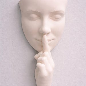 May include: A cream-coloured wall sculpture of a human face with closed eyes and a finger pressed to the lips, signalling silence. The sculpture is mounted on a light grey textured wall, creating a minimalist aesthetic.
