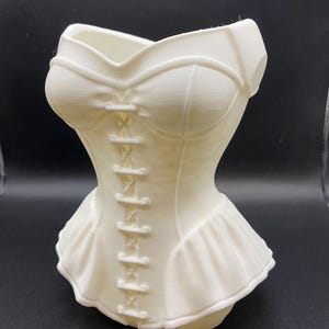 May include: A white, corset-shaped vase with a ruffled skirt detail. The vase features a detailed bodice with a lace-up design and structured cups. The overall design is a decorative, feminine piece, perfect for displaying flowers or as a standalone art object.