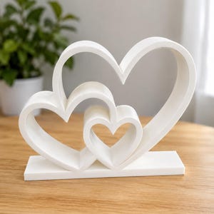 May include: A white decorative sculpture featuring three nested heart-shaped outlines of varying sizes, all connected to a single rectangular base. The object is constructed with smooth, matte surfaces and clean, continuous lines forming the heart shapes.