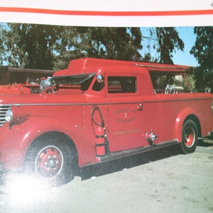 May include: A vintage red fire engine with a white cab and a wooden bed. The engine has a ladder on the side and a hose reel on the back. The engine is parked in front of a building with trees in the background.