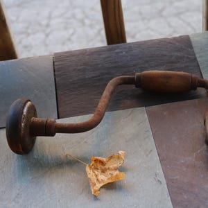 May include: An antique hand drill with a wooden handle and a rusty metal frame. The drill is lying on a slate surface next to a dried leaf. The tool is a vintage item.