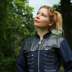 May include: A navy blue jacket with a black faux leather yoke and silver-toned hardware. The jacket features a zipper closure and decorative silver accents. The model is wearing white pants. The jacket is a unique fashion statement.