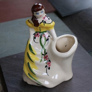 May include: Vintage ceramic figurine of a woman in a long dress, holding a basket. The dress is cream-colored with yellow accents and green and pink floral details. The woman has brown hair and blue eyes. A decorative piece.