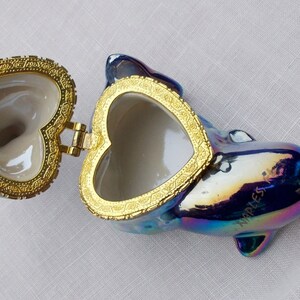May include: A iridescent blue dolphin shaped trinket box with a gold heart shaped lid. The lid is open and the inside of the box is white.
