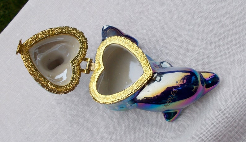May include: A iridescent blue dolphin shaped trinket box with a gold heart shaped lid. The lid is open and the inside of the box is white.