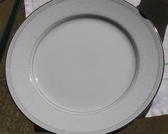 Può includere: Un piatto da portata in ceramica bianca con un bordo decorativo argento e bianco. Il piatto ha un bordo smerlato.