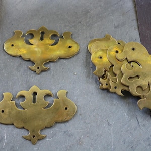 Op de afbeelding: Diverse antieke messing lade-knoppen met sleutelgaten en decoratieve ontwerpen. Het messing is verouderd en vertoont diverse goudtinten. Geschikt voor meubelrestauratie of knutselprojecten. Afmetingen in centimeters.