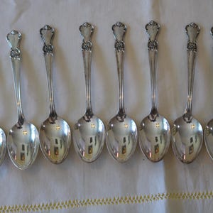 May include: A set of eight silver spoons arranged in a row. Each spoon has a rounded bowl and a long, slender handle. The handles are topped with a decorative floral design. The spoons are displayed on a white surface.