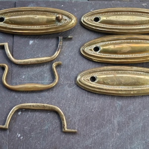 Op de afbeelding: Een assortiment antieke messing hardware, waaronder ovale achterplaten met decoratieve details en verschillende handgreepstijlen. De hardware is gerangschikt op een donkere, getextureerde ondergrond, wat de verouderde patina laat zien.