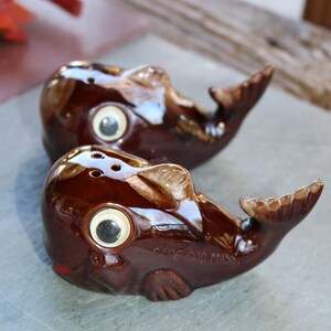May include: Two brown ceramic whale salt and pepper shakers with white eyes and black pupils. The shaker on the bottom has the words "Cape Cod Mass." written on the side.