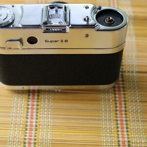 Pu&ograve; includere: Una macchina fotografica argentata e nera con il testo "Super II B" sul davanti. La macchina fotografica ha un rivestimento in pelle sul retro.