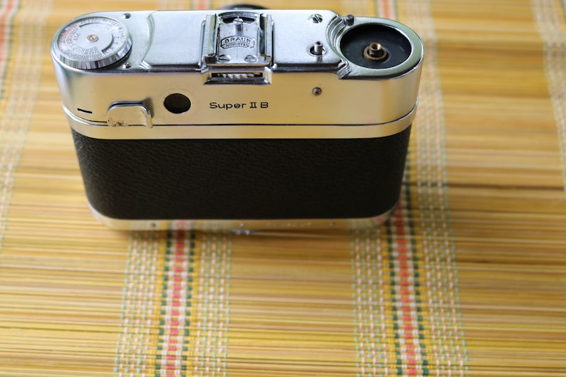 Pu&ograve; includere: Una macchina fotografica argentata e nera con il testo "Super II B" sul davanti. La macchina fotografica ha un rivestimento in pelle sul retro.