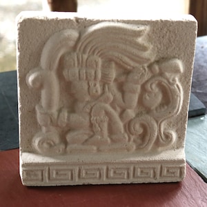 May include: A square, off-white stone tile featuring a relief carving of a seated figure wearing an elaborate headdress. The base of the tile is decorated with a repeating geometric Greek key pattern, and the surface has a matte, textured finish.