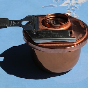 May include: A copper coffee pot with a stainless steel lid and a black handle. The lid has a small opening for pouring and a lever for opening and closing the lid. The pot is sitting on a blue surface.
