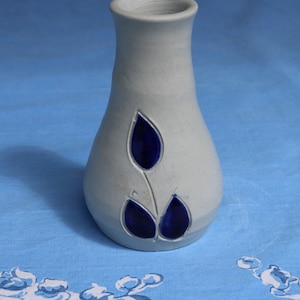 Puede incluir: Un jarrón de cerámica gris claro con la parte superior ensanchada y un cuerpo redondeado. El jarrón presenta un diseño de hojas azul oscuro. El jarrón está sobre un mantel azul claro con estampado floral.