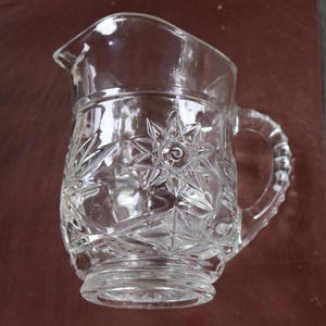May include: A clear glass pitcher with a starburst and geometric pattern. The pitcher has a handle and a spout for pouring. The glass is transparent, allowing light to pass through, and is likely used for serving beverages.