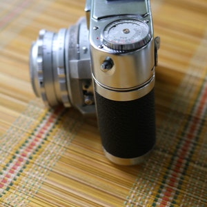 Pu&ograve; includere: Una macchina fotografica a pellicola da 35 mm argentata e nera con una impugnatura in pelle. La macchina fotografica ha un esposimetro incorporato e un obiettivo a messa a fuoco manuale.